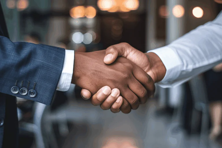 dois homens apertando as mãos é a foto que ilustra artigo sobre automatização de processos de vendas. Um está vestindo um terno azul e o outro uma camisa branca, com um fundo desfocado de ambiente corporativo.
