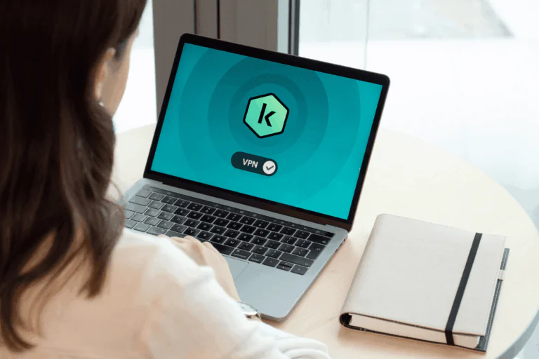 ilustrando artigo sobre Kaspersky VPN, uma mulher está sentada de costas, utilizando um notebook em uma mesa redonda. Na tela do notebook, é exibido um logotipo com a letra "K", logo da Kaspersky, e o texto "VPN" com um símbolo de verificação ao lado, indicando que a conexão VPN está ativa. Ao lado do notebook, sobre a mesa, há um caderno de capa clara.