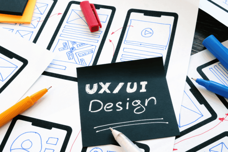 Imagem de rascunhos de design e melhores práticas de UX/UI para aplicativos móveis. No centro, um cartão preto com a inscrição 'UX/UI Design' em letras brancas está rodeado por desenhos esquemáticos de interfaces móveis feitas em papel, com setas e anotações indicando o fluxo de navegação. Canetas coloridas e materiais de escritório complementam a cena, representando o processo criativo de desenvolvimento de design de interface e experiência do usuário.