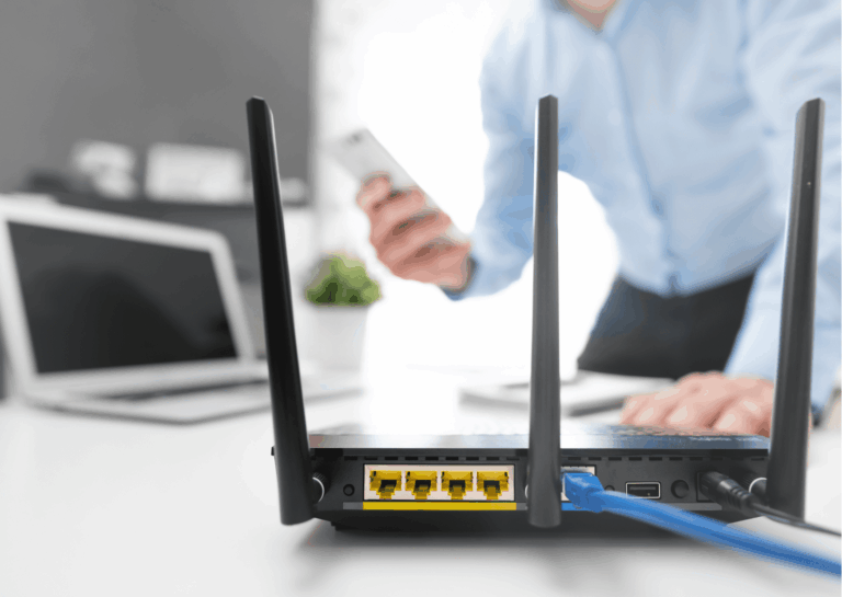 ao fundo, em fotografia desfocada, mesa de escritório com laptop desligado, homem segurando aparelho de celular e a outra mão sobre a mesa; em destaque, no centro e canto direito, um aparelho roteador com três antenas externas; exemplificando como configurar a rede wireless no Windows.