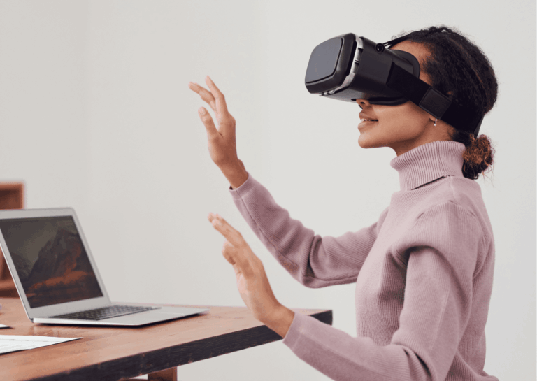 mulher negra, vestindo suéter lilás de gola alta, está sentada e usando um óculos de realidade virtual aumentada, com as mãos espalmadas para frente e sorrindo; à sua frente, uma mesa de madeira com um laptop prateado aberto com uma paisagem de montanha na tela; demonstra como preparar sua empresa para o Metaverso.