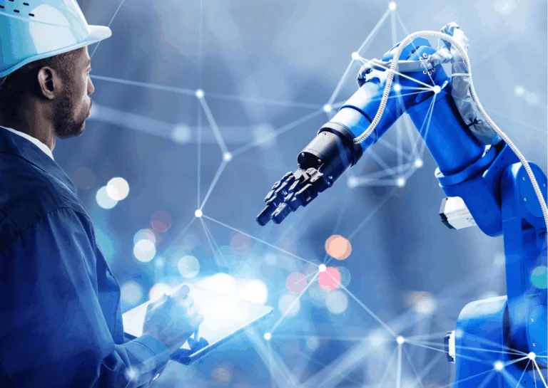 Um braço mecânico está estendido do canto inferior esquerdo da imagem até o centro e, do lado direito, um homem negro usa um jaleco azul, capacete e segura uma prancheta; exemplificando o que é automação industrial e robótica.