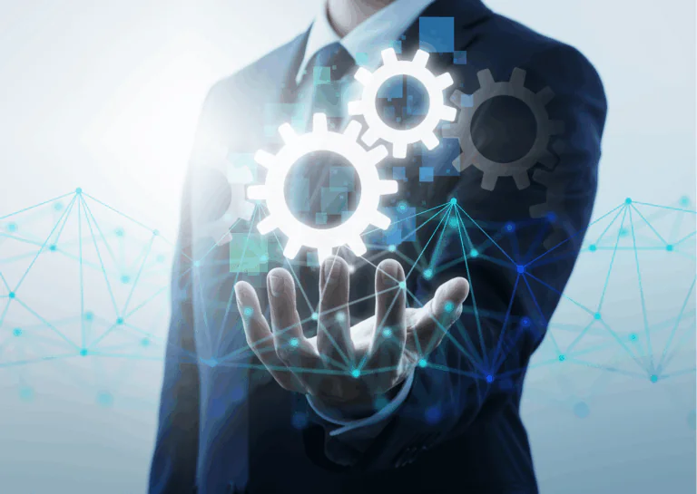 Sobre fundo azul claro, um homem vestido de terno escuro, que aparece apenas seu torço, está com a mão esquerda estendida para a frente, com a palma virada para cima e, sobre ela, estão desenhadas digitalmente engrenagens dispostas umas interligadas às outras, e, abaixo e por cima da mão, de uma ponta a outra da imagem, uma rede de fios com pontos que os conectam, em azul, se estende; exemplifica a importância da automatização de processos e tarefas.