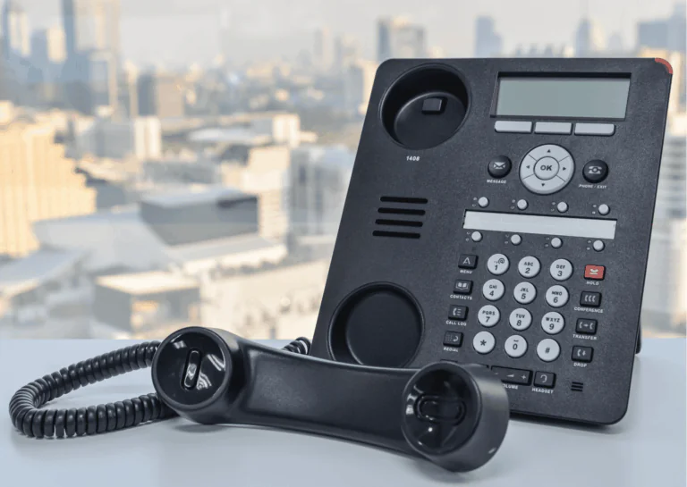 Sobre superfície plana e branca, está um aparelho telefônico do tipo PABX, com o fone fora do gancho, deitado na frente do aparelho; ao fundo, a vista desfocada de uma cidade; exemplifica o que é um telefone IP.