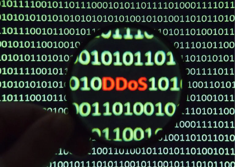 Representando o que é um ataque DDos, a imagem mostra sobre um fundo preto, linhas de códigos binários em cor verde, e, sobre ela, aparece a parte de uma mão segurando uma lupa, que dá ênfase ao escrito DDoS, na cor vermelha, no meio de uma das linhas de numerais zero e um.