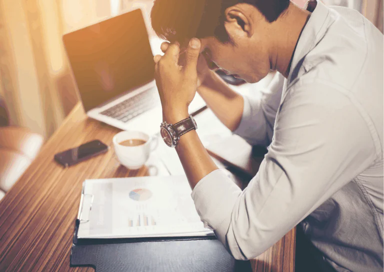 Sentado em frente à uma mesa de trabalho, com notebook, xícara de café e pastas com papéis e gráficos, está um homem usando camisa social, com os cotovelos apoiados na mesa e as mãos apoiando a cabeça, com os dedos nas têmporas; representado como as empresas podem contribuir para evitar o burnout.