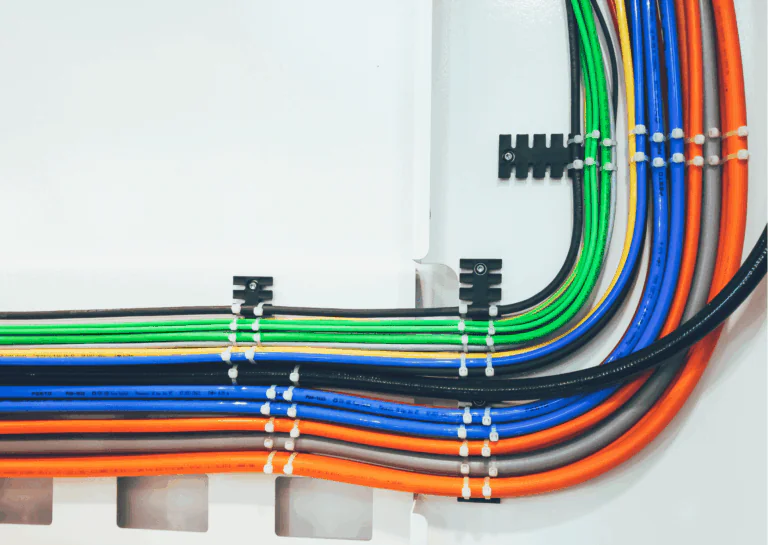 Representando o conceito e a importância do cabeamento estruturado, está em destaque uma série de fios nas cores verde, amarelo, cinza, azul, preto e alaranjado, organizados e presos por canaletas e conduítes, sobre uma superfície branca.