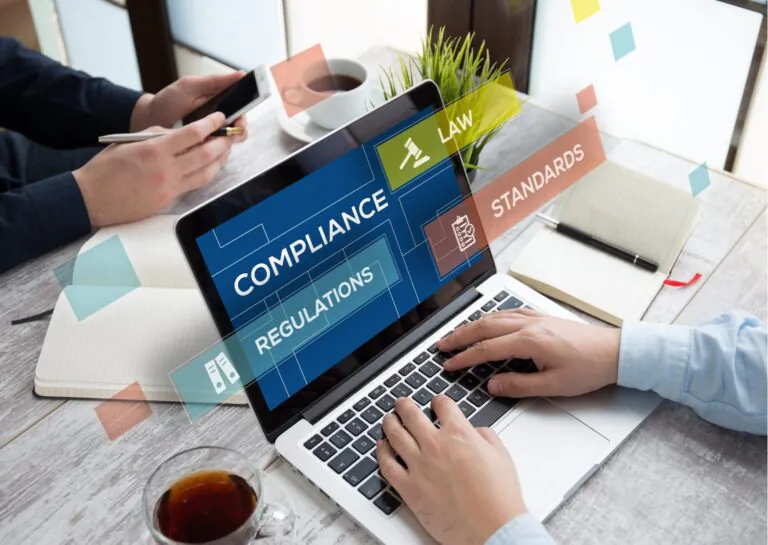 A fotografia exemplificativa do papel do compliance na gestão dos negócios mostra sobre uma musa um notebook em foco, com a tela mostrando a palavra compliance em fundo azul e em retângulos saltados da tela as palavras regulations, law e standards; ao redor na mesa aparece os braços de uma pessoa com mangas compridas azul clara, dispostas sobre o teclado, cadernos ao lado e uma xícara transparente de vidro com café e, ao fundo os braços de outra pessoa segurando um smartphone e uma caneta, sobre um caderno aberto, com xícara e uma planta pequena ao lado, também sobre a mesa.