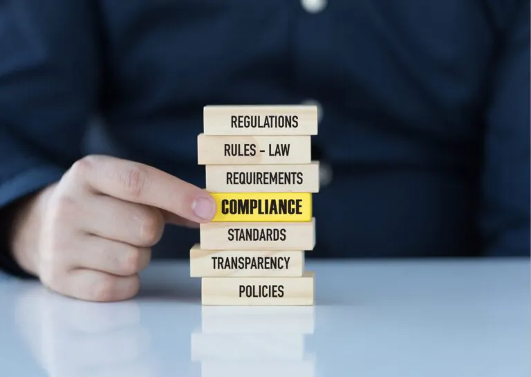 Representando o que é controle interno, a fotografia mostra uma pessoa sentada atrás de uma mesa, em desfoque ao fundo, aparecendo apenas parte do torso, e, em foco, em cima da mesa, estão blocos de madeira com escritos em preto: regulations, rules - law, requirements, compliance, standards, transparency, policies, sendo que o bloco em que está escrito compliance está no meio da pilha, é amarelo e a mão da pessoa está com indicador e polegar o segurando pela ponta.