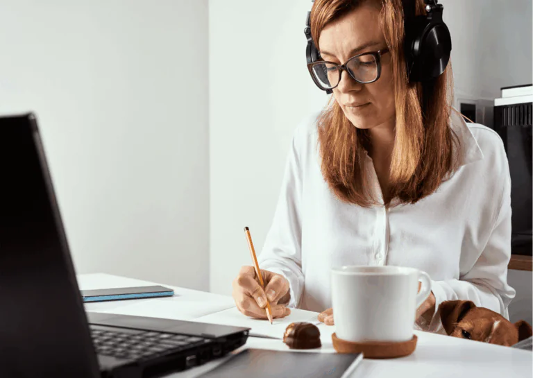 Sentada em frente à uma mesa com um notebook aberto à frente, uma mulher veste camisa branca, usa óculos de grau e um headphone, está fazendo anotações a lápis em um papel, com uma caneca branca ao lado, celular e papéis; exemplifica os cursos gratuitos para Office 365.