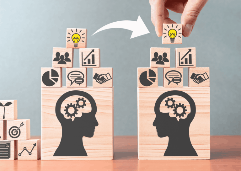 Sobre um superfície plana, de madeira, atrás um fundo cinza, estão dispostos blocos de madeira clara, empilhados uns nos outros, com desenhos na cor cinza estampados e representando pessoas, gráficos, lâmpada e balões de conversa; acima dos blocos, uma mão humana segura um deles que tem o desenho de lâmpada, para o qual uma seta branca saindo de outro bloco com lâmpada desenhada, também aponta; representa o que é PmBok.