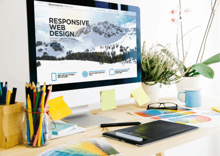 Sobre uma mesa de escritório, colocada rente à uma parede branca, está um desktop branco com borda da tela preta e, na tela, aparece a imagem de uma montanha, aparentemente um site de teste, com título em destaque escrito "responsive web design"; na frente do desktop e ao redor, estão espalhados materiais de escritório, como porta-lápis, prancheta, papéis, óculos, caneca, post-its na tela, cadernetas, caneta, etc., e, ao fundo e canto direito, algumas plantas e orquídeas; exemplificam o que é um plugin.