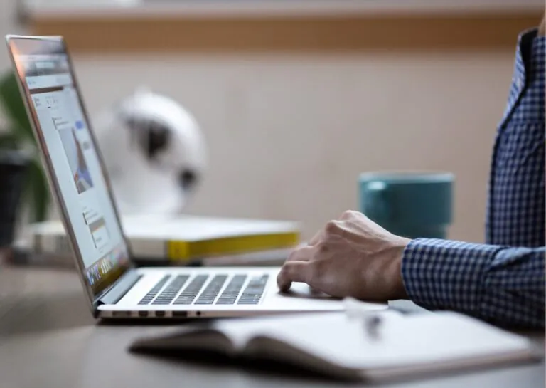 Representando o que é a Power Platform, em um ambiente de escritório, a fotografia mostra em destaque um notebook sobre uma mesa, com apenas os braços de uma pessoa branca sobre a mesa e teclado usando camisa de manga comprida azul e xadrez, com fundo desfocado mostrando livro, caneca, globo terrestre e agenda aberta.