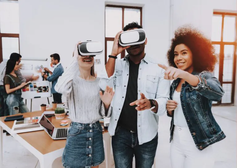 Representando os benefícios da realidade virtual para empresas, a fotografia mostra um ambiente de trabalho com mesa ao fundo e uma equipe trabalhando em um moodboard, e três pessoas em destaque à frente, duas delas utilizando óculos de realidade virtual e uma delas sem óculos, explicando seu uso.