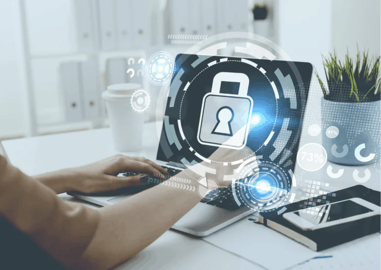 Representando a segurança digital para empreendedores, está uma fotografia de um ambiente de trabalho com mesa branca onde estão dispostos notebook, copo branco de café, agenda e celular e aparece parte de braços de uma mulher que está com as mãos sobre o laptop e com o indicador da mão direita tocando a tela do aparelho; sobre esta imagem, está um círculo formado por várias partes como uma engrenagem sem dentes e, no centro, está o desenho de um cadeado branco.
