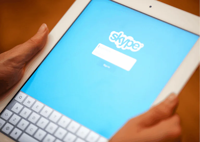 Em foco na imagem, aparecem duas mãos segurando um tablet branco que em sua tela aparece a imagem de login do Skype, azul com o nome da marca e espaço para digitação do login, abaixo, o teclado está ativado na tela do tablet; exemplifica as vantagens do Skype para empresas.