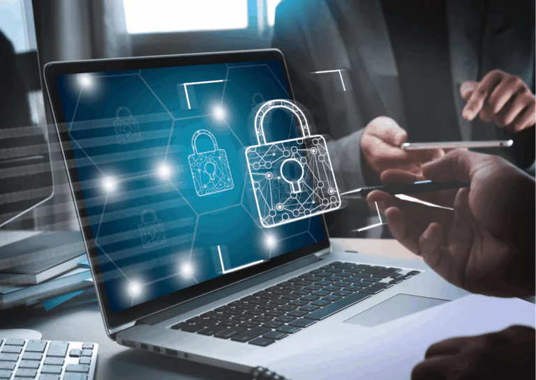 Em destaque está um notebook na diagonal, em cima de uma mesa, com a tela azul mostrando alguns desenhos de cadeados na cor branca; flutuando à frente da tela está a imagem de cadeado, também branco, e, ao redor, é possível ver partes de duas pessoas, uma sentada ao fundo, segurando um celular na posição horizontal, e com a mão em posição de tocar na tela e a outra pessoa, da qual aparecem apenas uma mão direita segurando uma caneta e uma parte da mão esquerda sobreposta em frente ao notebook; estão representando as tendências de segurança cibernética para 2023.